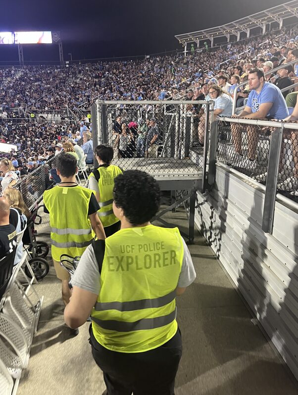 Three explorers wearing yellow vests walking through the stands at a UCF football game.