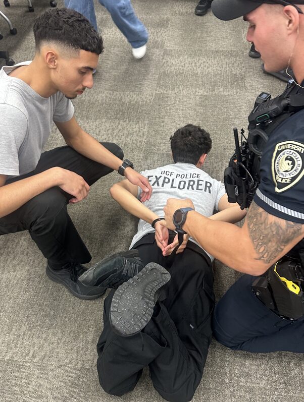 A police officer and an explorer demonstrating a suspect apprehension technique. Suspect is on the floor with feet and hands in handcuffs.